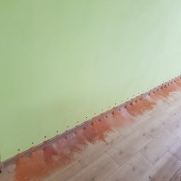 MAD-MAX Sp. z o.o. - Dolna część ściany w pokoju z widocznymi śladami wilgoci i pleśni oraz otworami wentylacyjnymi wzdłuż podłogi, sugerujące problem z wilgocią