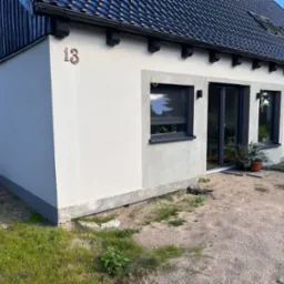 Wykończony budynek z numerem '13', z czarną dachówką, oknami i drzwiami przesuwnymi. Widoczne świeże tynki i szara podmurówka. Otoczenie to piasek i trawa.