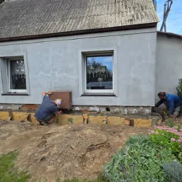 Dwóch robotników przy izolacji fundamentów domu jednorodzinnego. Widoczne drewniane szalunki i świeży beton. Praca w toku, zieleń w tle.