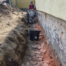 MAD-MAX Sp. z o.o. - Prace przy izolacji fundamentów: pracownik z maszyną w wykopie wzdłuż ściany budynku, widoczne cegły i wiadro. Etap wiercenia otworów pod iniekcję.