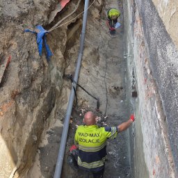 MAD-MAX Sp. z o.o. - Pracownicy firmy MAD-MAX IZOLACJE w głębokim wykopie przy fundamencie budynku, wykonujący prace izolacyjne. Widoczna odsłonięta ceglana ściana i szara rura drenażowa.