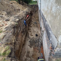 MAD-MAX Sp. z o.o. - Głęboki wykop wzdłuż ściany budynku z widocznym fundamentem i warstwą izolacyjną. Pracownik w kamizelce pochylony w dole, obok drabina i narzędzia.