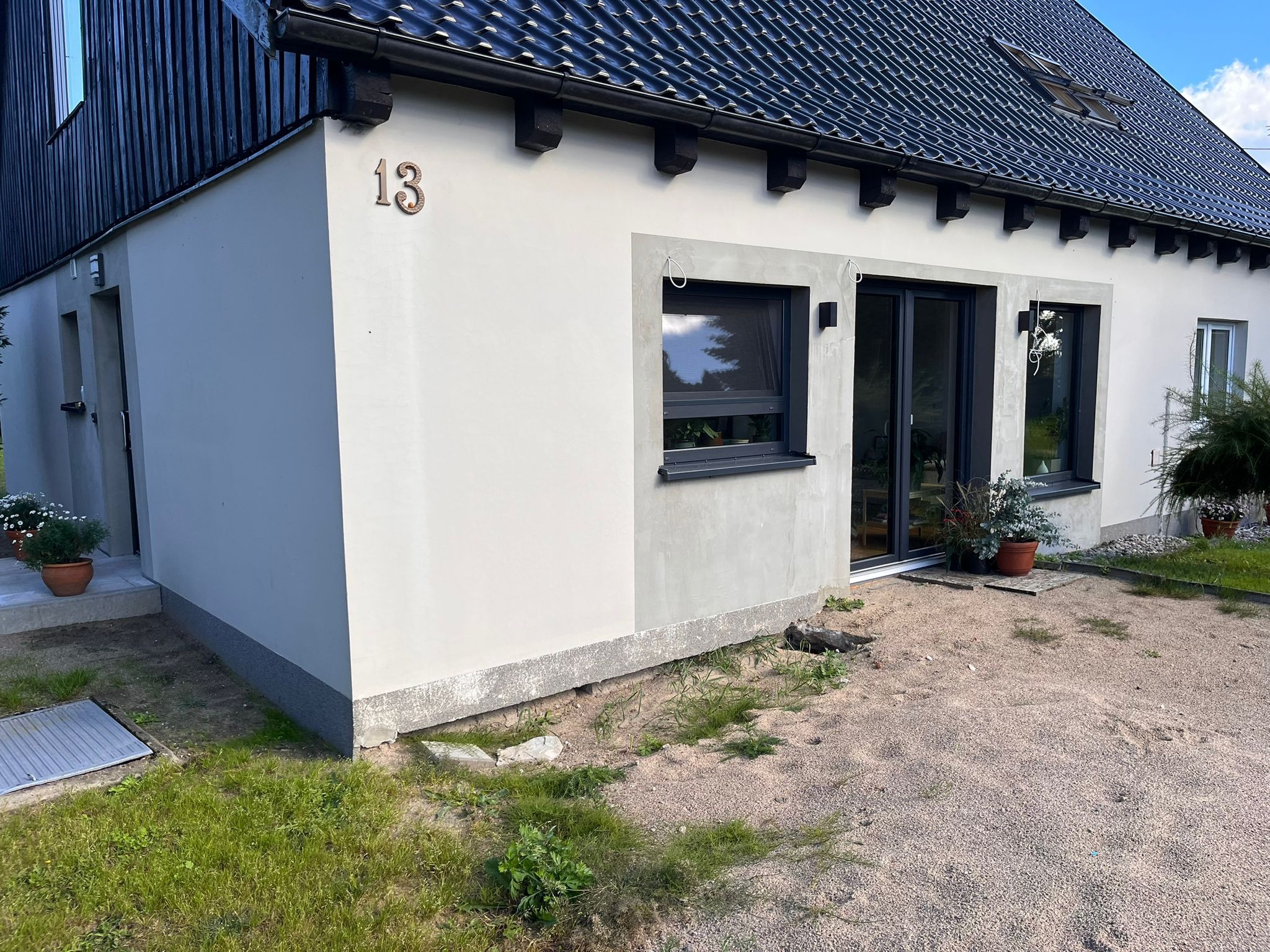 Wykończony budynek z numerem '13', z czarną dachówką, oknami i drzwiami przesuwnymi. Widoczne świeże tynki i szara podmurówka. Otoczenie to piasek i trawa.