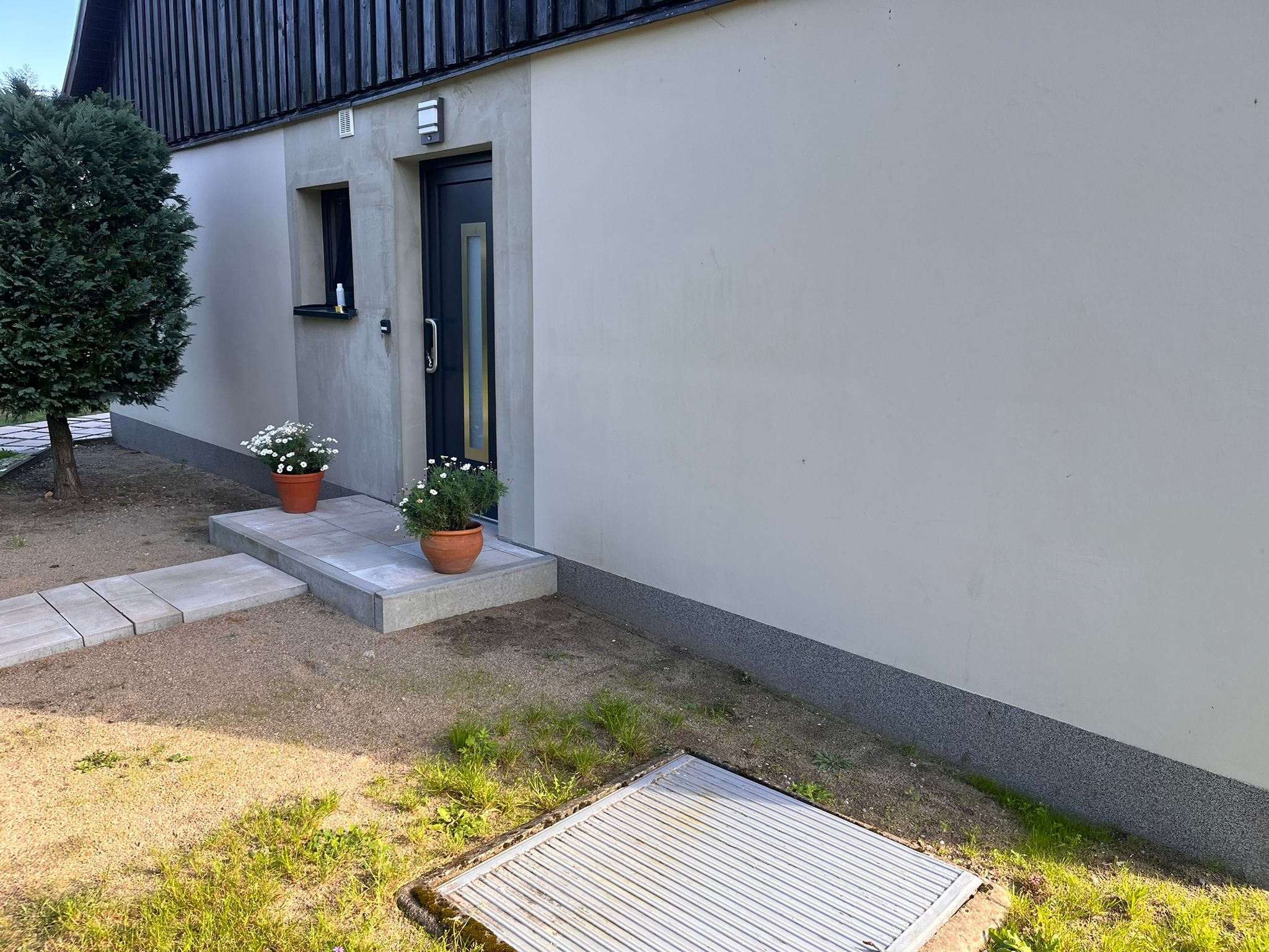 Elewacja domu po remoncie: jasny tynk, ciemne okna i drzwi. Dwa kwiaty w donicach przy wejściu, schludny chodnik i trawnik. Widoczny fragment dachu.