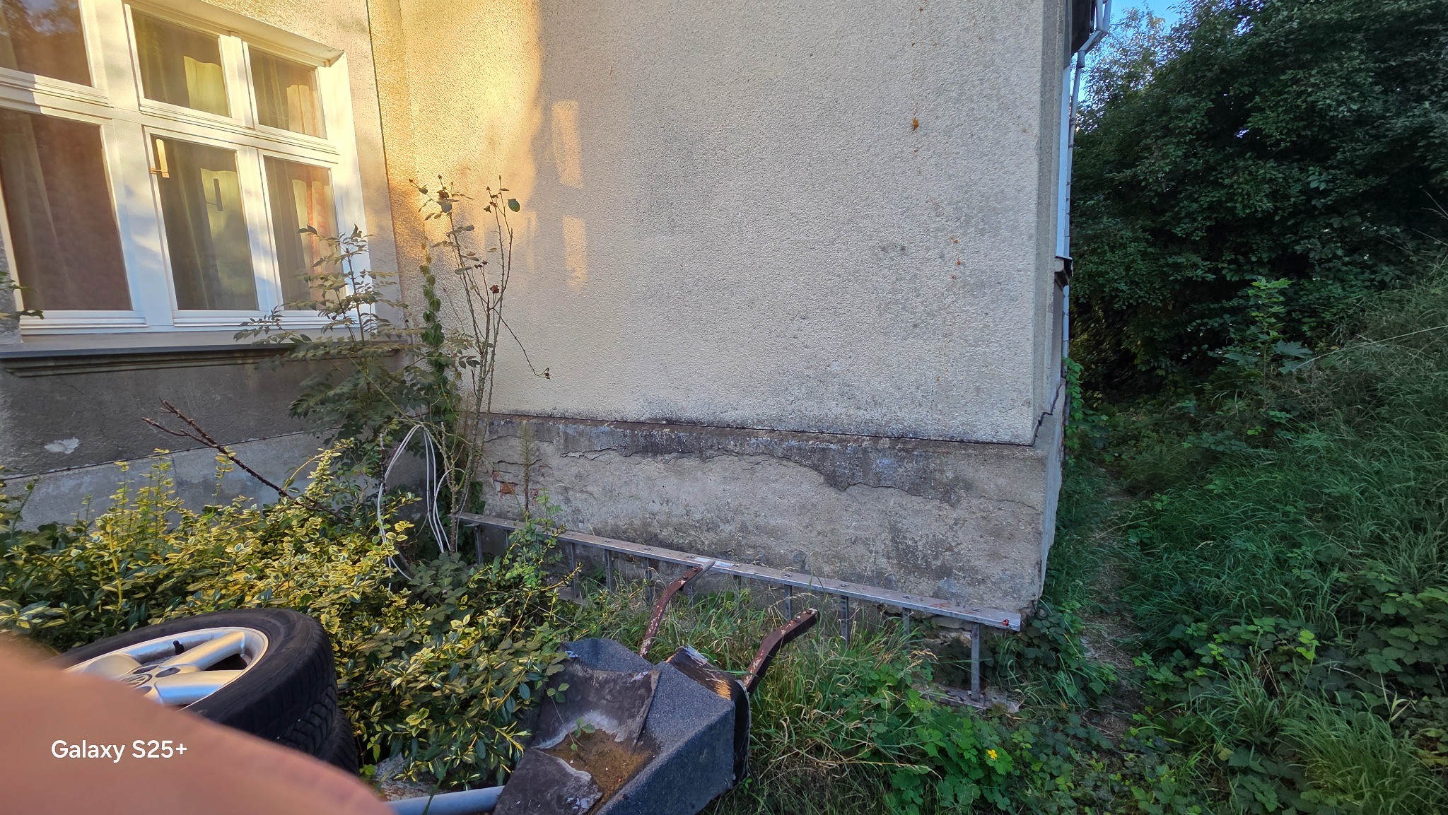 Uszkodzona elewacja budynku z widocznymi pęknięciami przy fundamencie, zarośnięta roślinnością, z fragmentem okna i opony w kadrze. Widoczne ślady wilgoci.