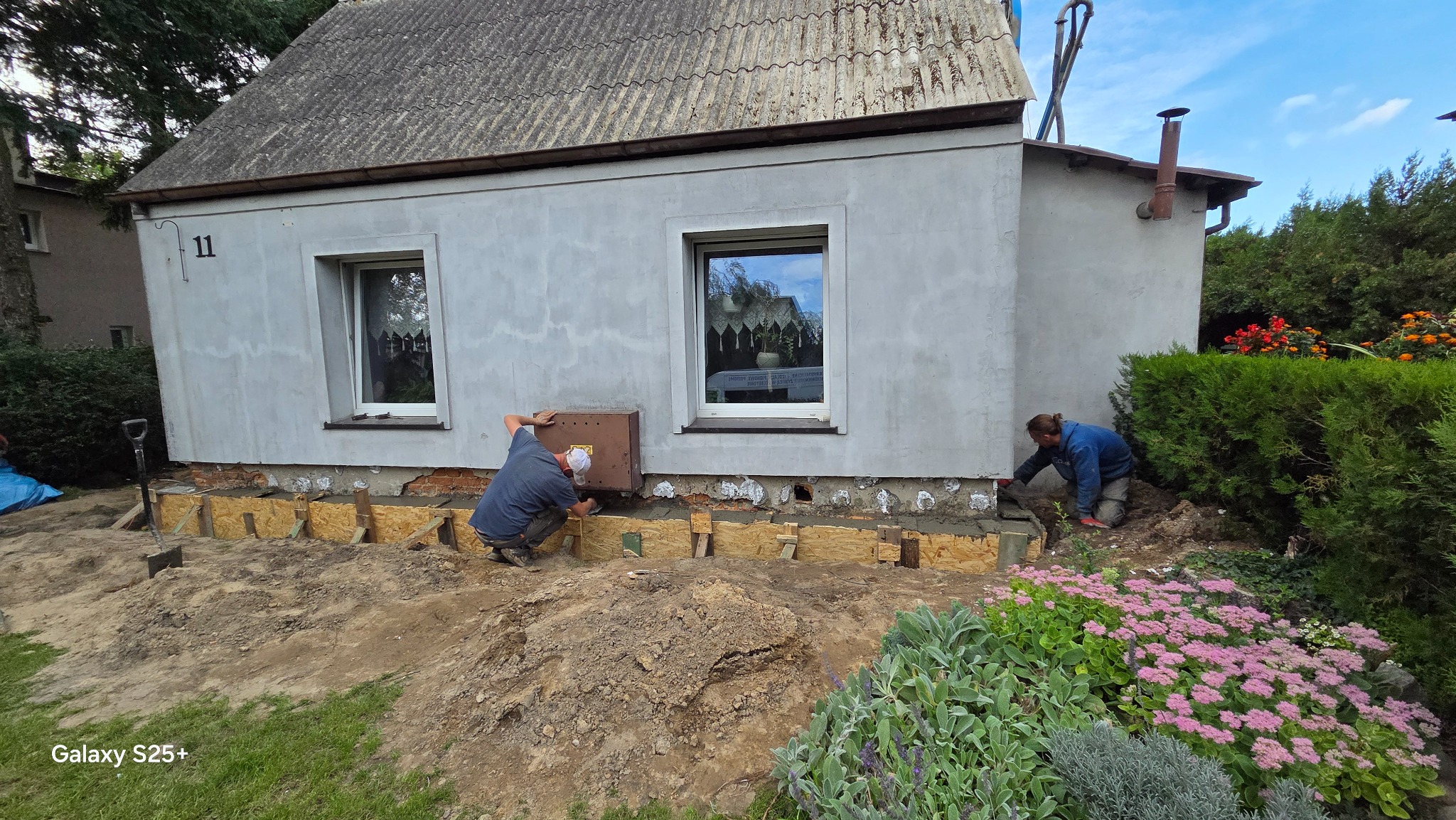 Dwóch robotników przy izolacji fundamentów domu jednorodzinnego. Widoczne drewniane szalunki i świeży beton. Praca w toku, zieleń w tle.