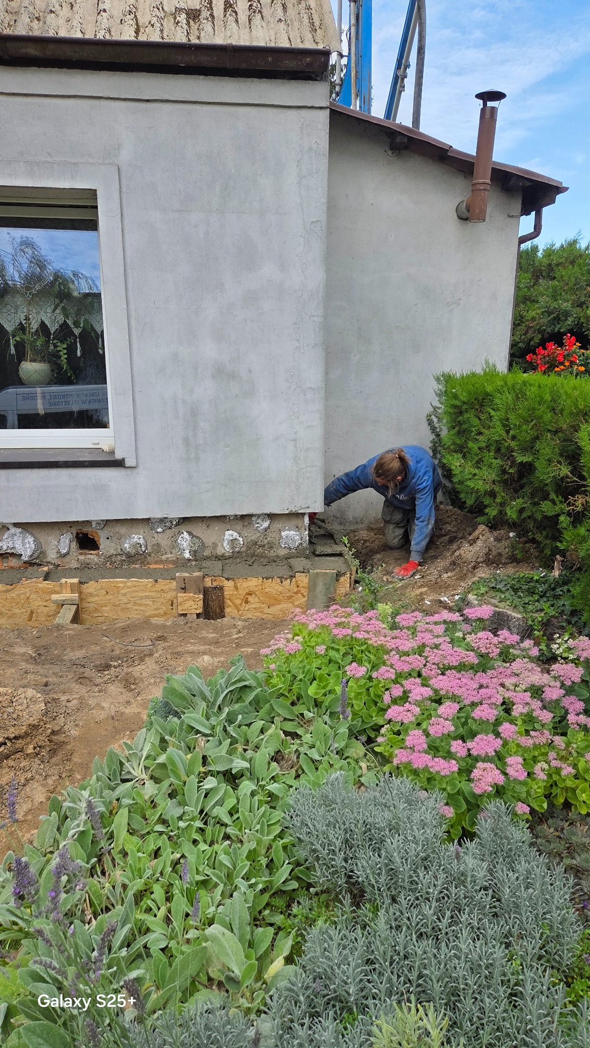 Pracownik w trakcie izolacji fundamentów domu jednorodzinnego, widoczne szalunki i częściowo wykonana izolacja. W tle roślinność ogrodowa i fragment betonomieszarki.