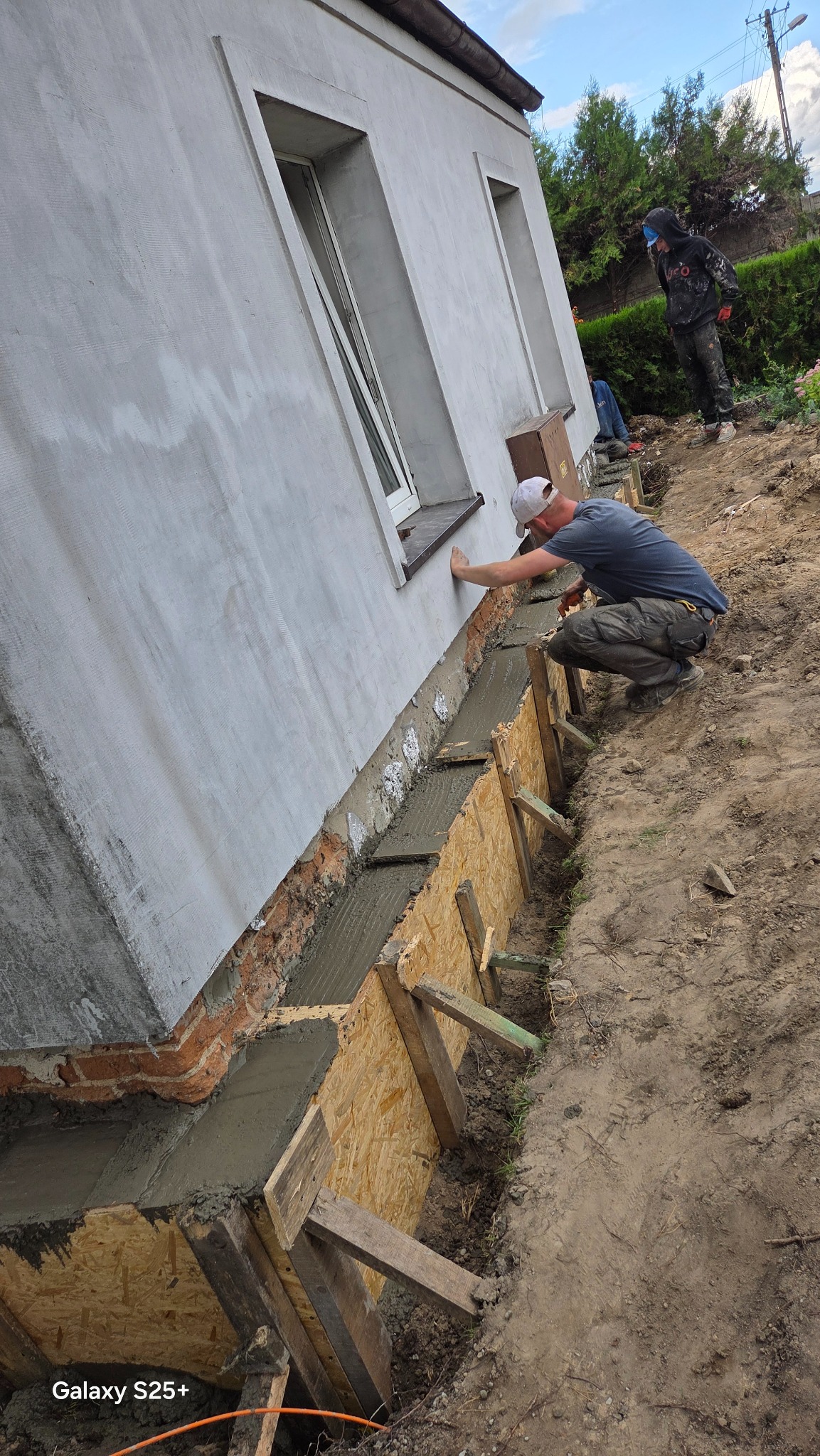 Pracownik w trakcie izolacji fundamentów budynku, widoczna drewniana forma zalana betonem, ściana budynku z oknami. Prace ziemne wokół budynku.