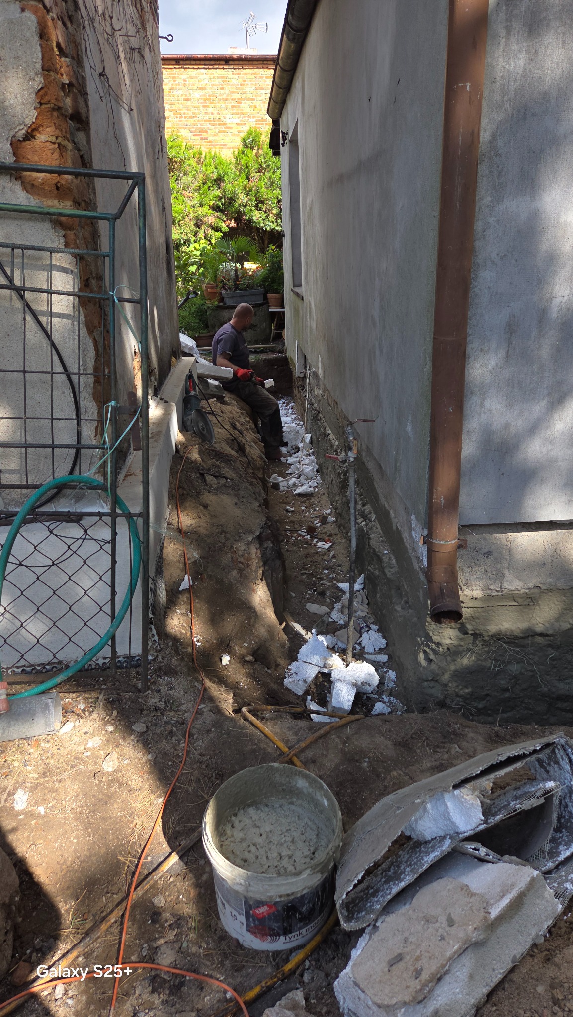 Prace ziemne przy budynku: fundament odsłonięty, widoczny pracownik z narzędziami, przygotowania do izolacji. W tle zieleń i ceglany budynek.