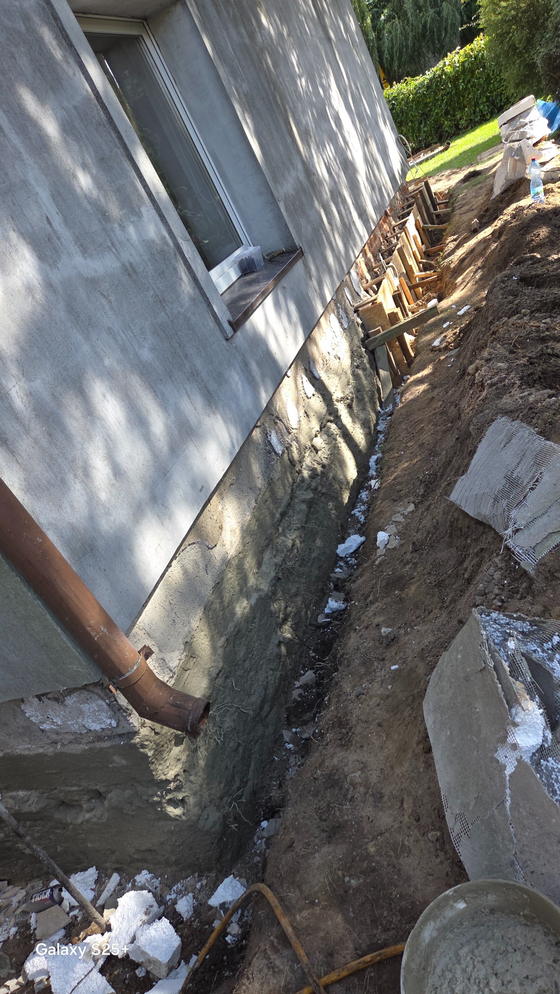 Fundament budynku w trakcie izolacji: wykop z deskowaniem, widoczny fragment ściany z rurą spustową. Prace ziemne w toku, elementy izolacyjne w wykopie.