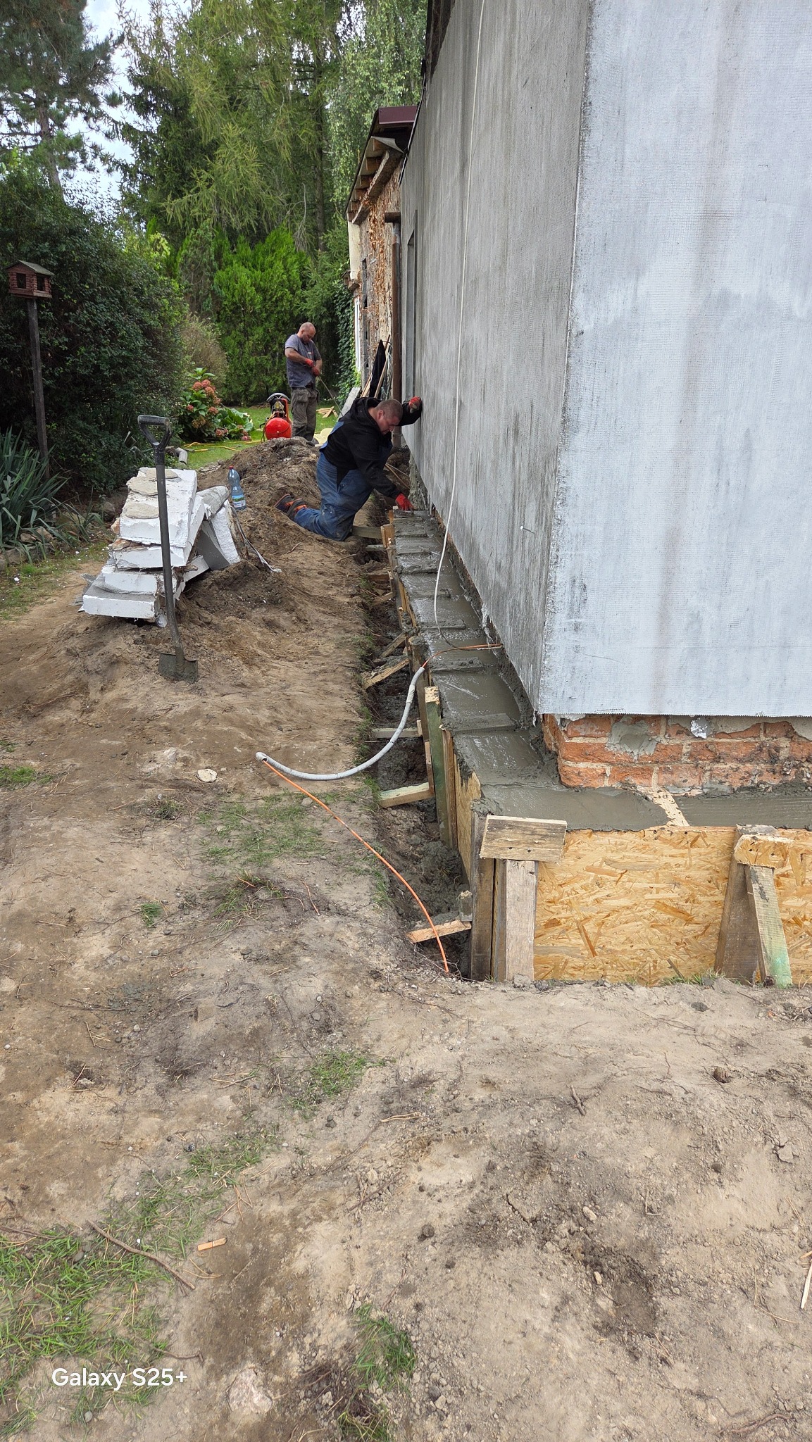 Prace przy izolacji fundamentów budynku: robotnik w czerwonych rękawicach układa beton w szalunku. Widoczny mur z cegły, drugi pracownik w tle. Przewód wentylacyjny.