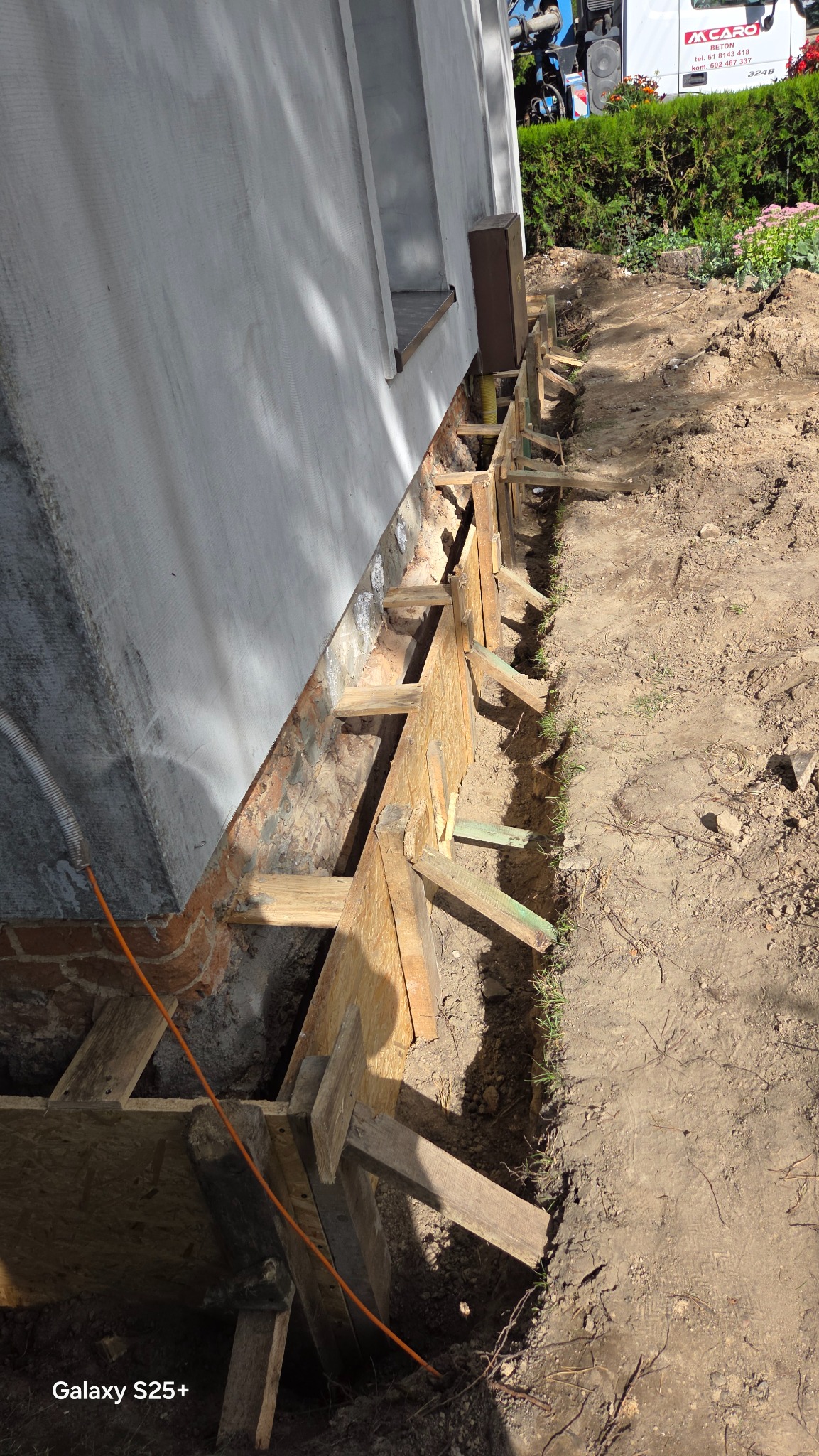Fundament budynku w trakcie izolacji, widoczne drewniane szalunki i wykop. Elementy instalacji wystają spod ściany. W tle betoniarka firmy MCARO.