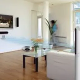 Salon z zamontowanym systemem nagłośnienia sali konferencyjnej: telewizor na ścianie otoczony czarnymi głośnikami, biała sofa i stolik kawowy z kwiatami, parkiet na podłodze.