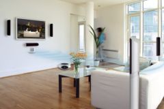 Salon z zamontowanym systemem nagłośnienia sali konferencyjnej: telewizor na ścianie otoczony czarnymi głośnikami, biała sofa i stolik kawowy z kwiatami, parkiet na podłodze.