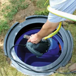 Montaż pompy zanurzeniowej w zbiorniku oczyszczalni ścieków. Widoczny fragment trawnika i dłoń instalatora wkładająca pompę do wnętrza zbiornika z niebieskim dnem.