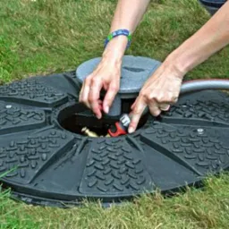 Czarna pokrywa studni na trawniku, z podłączonym wężem ogrodowym. Widoczne dłonie osoby obsługującej złącze.