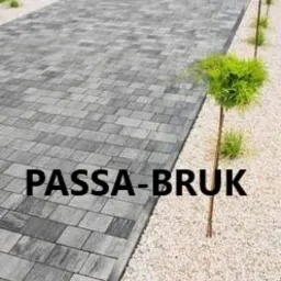 Szara kostka brukowa ułożona w zygzakowaty wzór, obok wyłożona drobnym białym żwirem, w tle małe drzewka ozdobne.