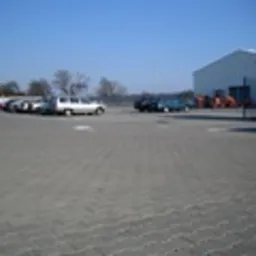 Rozległy plac wyłożony szarą kostką brukową, z widocznymi zaparkowanymi samochodami osobowymi i budynkiem magazynowym w tle, sfotografowany w słoneczny dzień.