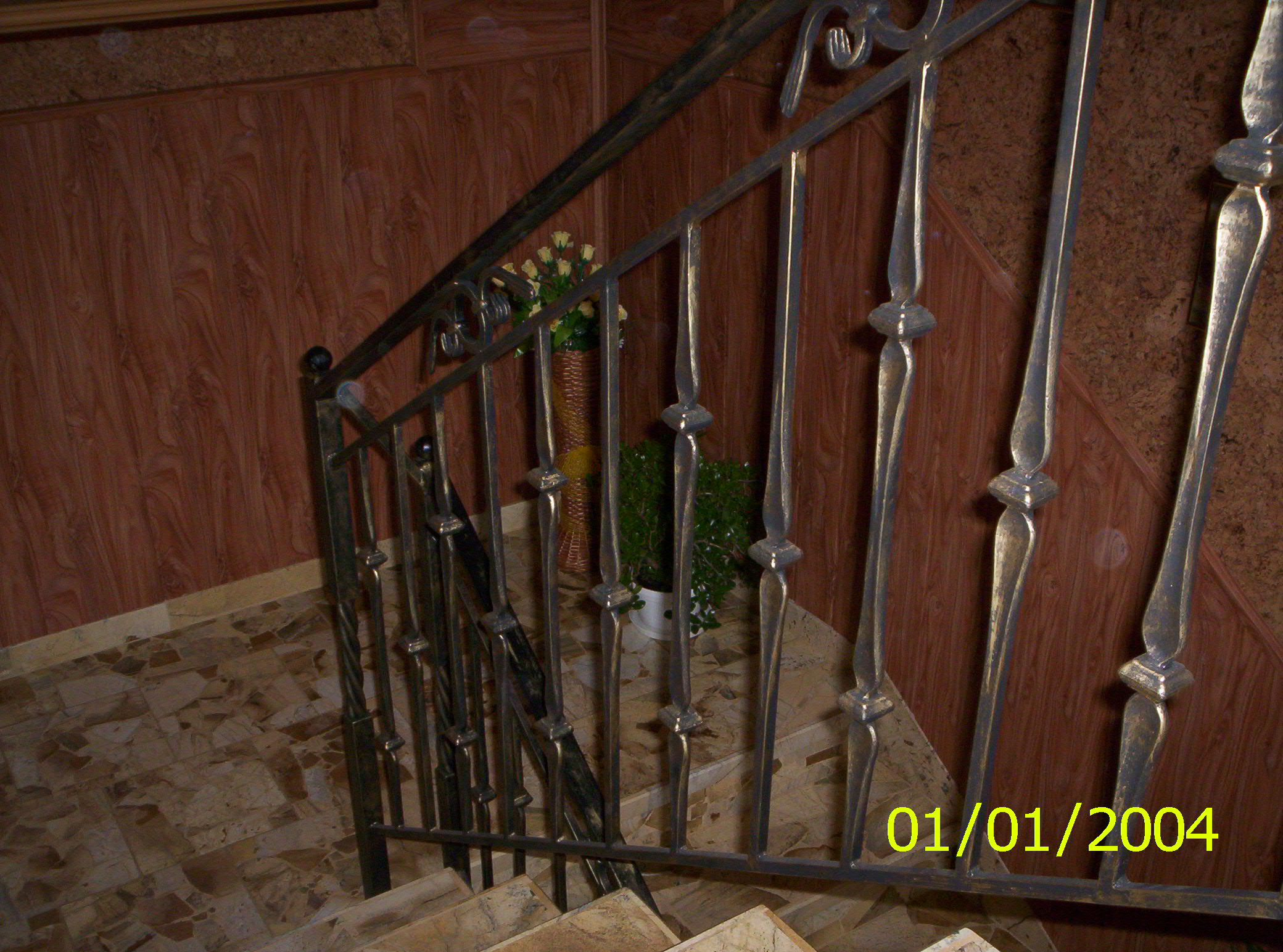 Kuta, metalowa balustrada schodowa z dekoracyjnymi elementami, widok z góry na schody wyłożone płytkami, w tle boazeria i kwiaty.