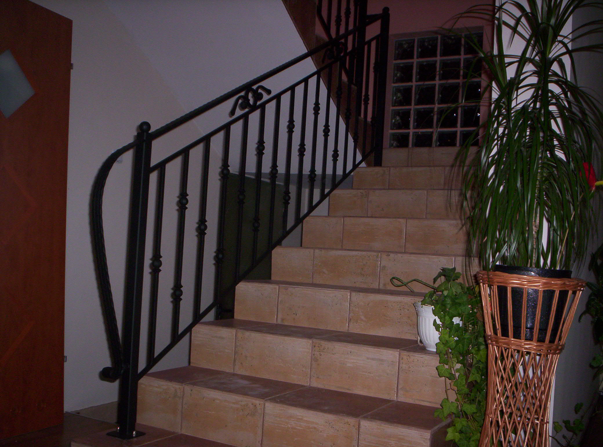 Kute, czarne balustrady schodowe z dekoracyjnymi elementami na stopniach wyłożonych beżowymi płytkami, obok doniczka z wysoką rośliną.