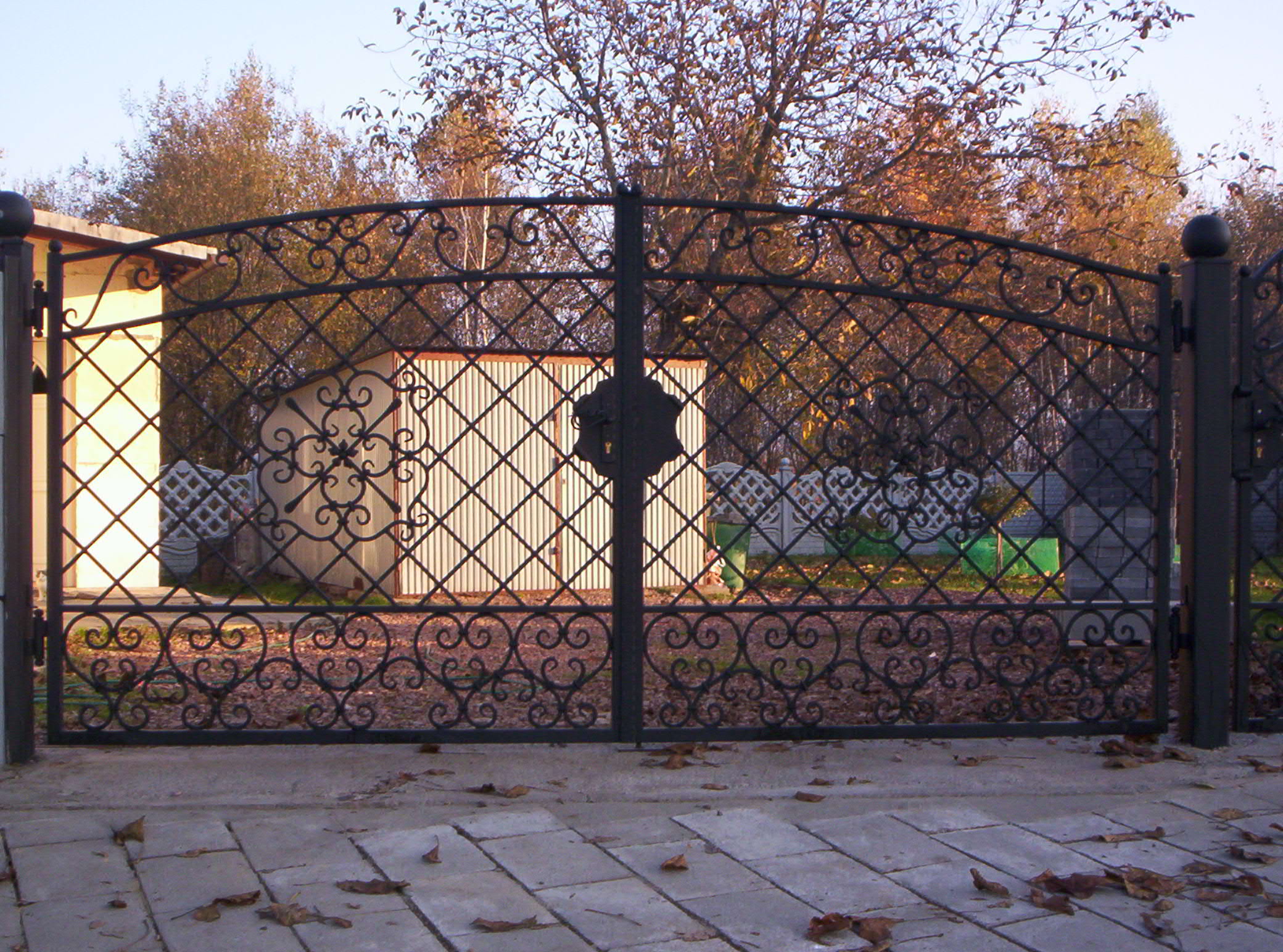 Kuta, metalowa brama ogrodzeniowa z dekoracyjnymi elementami w kształcie serc i rombów, widok z zewnątrz na podjazd wyłożony kostką, w tle budynek gospodarczy i drzewa.