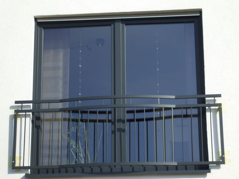 Szary metalowy balkon z prostymi pionowymi prętami i poziomymi poręczami, zamontowany przed dużym oknem na białej ścianie budynku, zabezpieczony taśmą malarską.