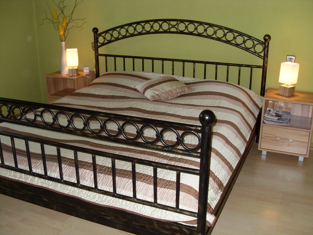 Kute metalowe łóżko z okrągłymi zdobieniami na ramie i wezgłowiu, przykryte kołdrą w brązowo-białe pasy, stoi w pokoju z zielonymi ścianami obok drewnianych szafek nocnych z lampkami.