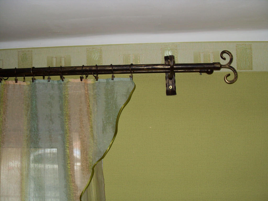 Kuty karnisz w kolorze ciemnego złota z ozdobnym zakończeniem w kształcie spirali, zamocowany na zielonej ścianie z delikatną tapetą, z zawieszoną, półprzezroczystą zasłoną w odcieniach zieleni...