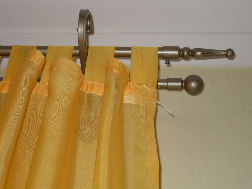 Żółte, prześwitujące zasłony zawieszone na metalowym karniszu z ozdobnymi zakończeniami w kształcie kuli i grotu.