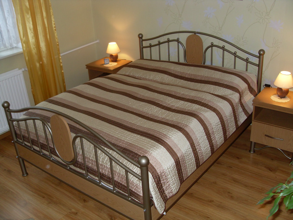Metalowe łóżko z drewnianymi elementami, nakryte brązowo-beżową kołdrą w paski, z dwiema lampkami nocnymi na szafkach po bokach, na drewnianej podłodze.
