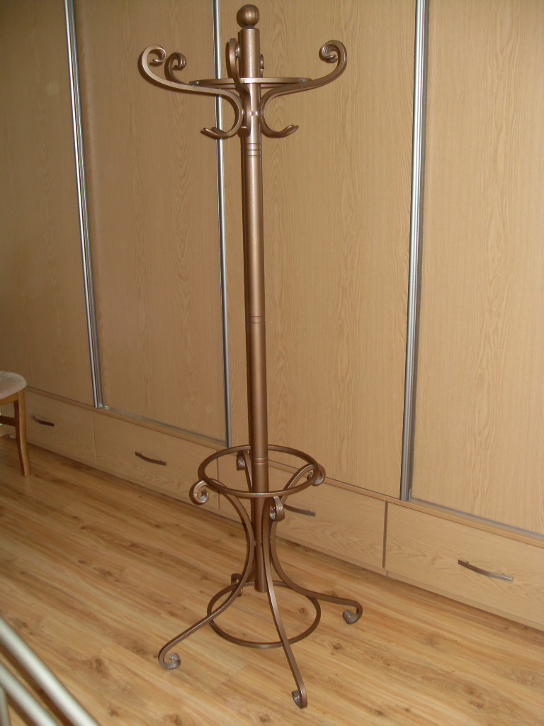 Kowalstwo artystyczne: Metalowy wieszak stojący w kolorze starego złota, z ozdobnymi zawijasami i kulką na szczycie, na tle drewnianej szafy przesuwnej.