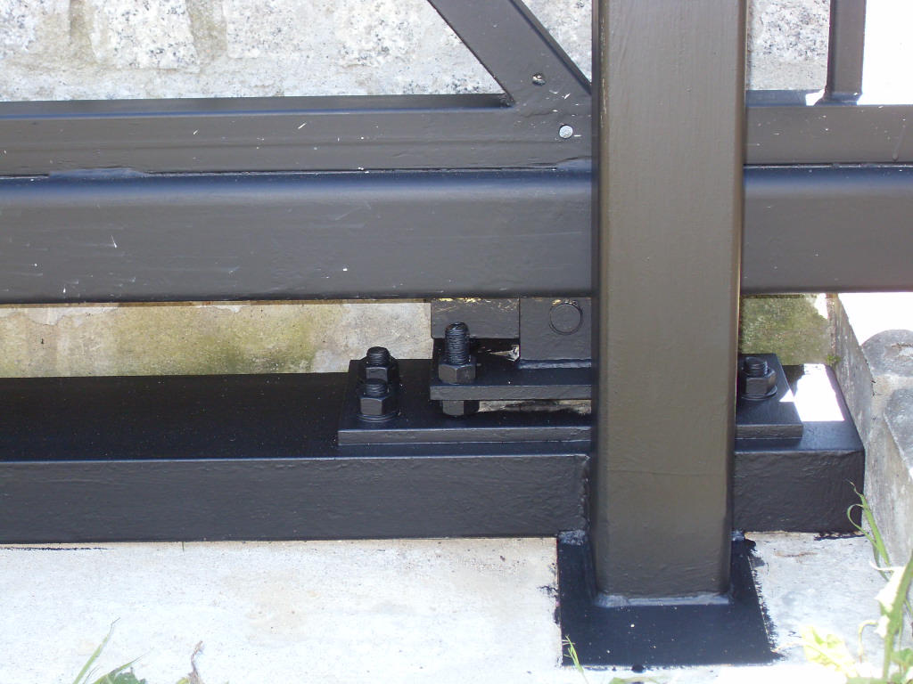 Szczegół mocowania czarnej, metalowej bramy do betonowego podłoża za pomocą śrub i nakrętek, widoczne ślady spawania i świeża farba.