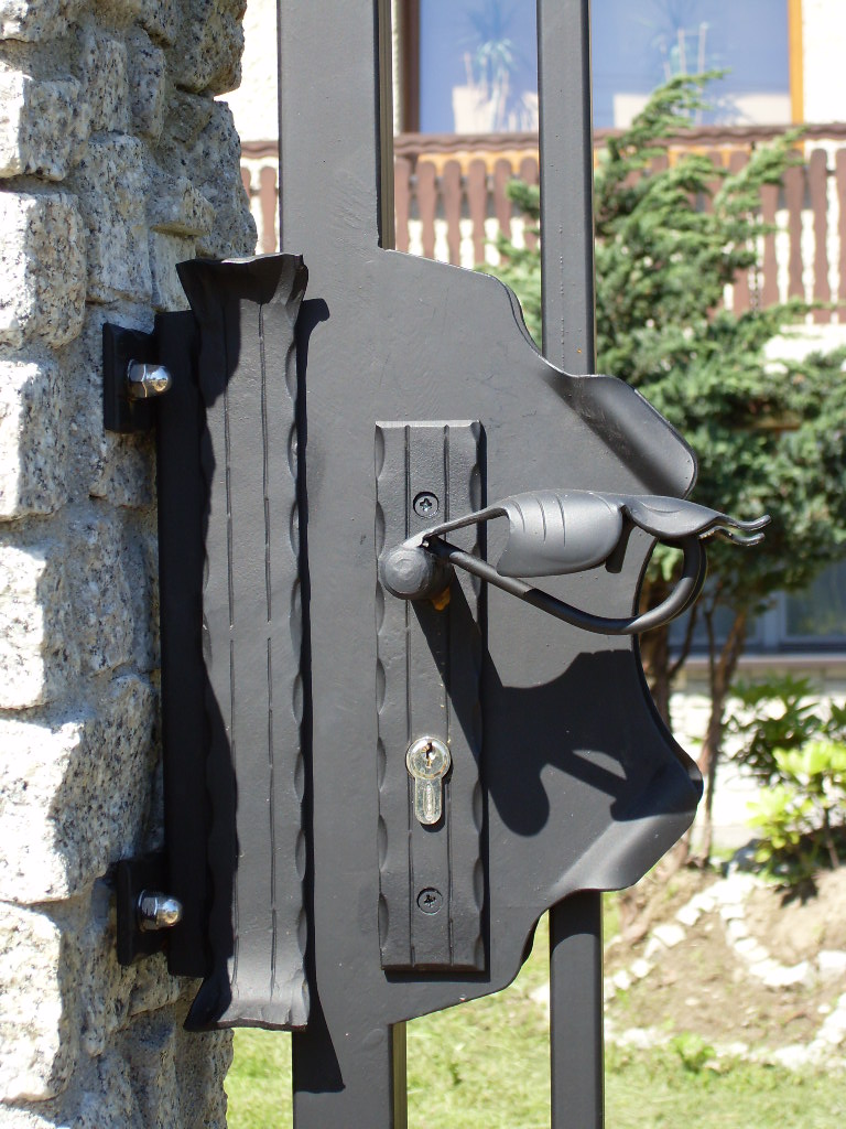 Czarny, metalowy element bramy z ozdobnym uchwytem w kształcie liścia, zamocowany do kamiennego słupka z widocznym zamkiem i śrubami mocującymi.