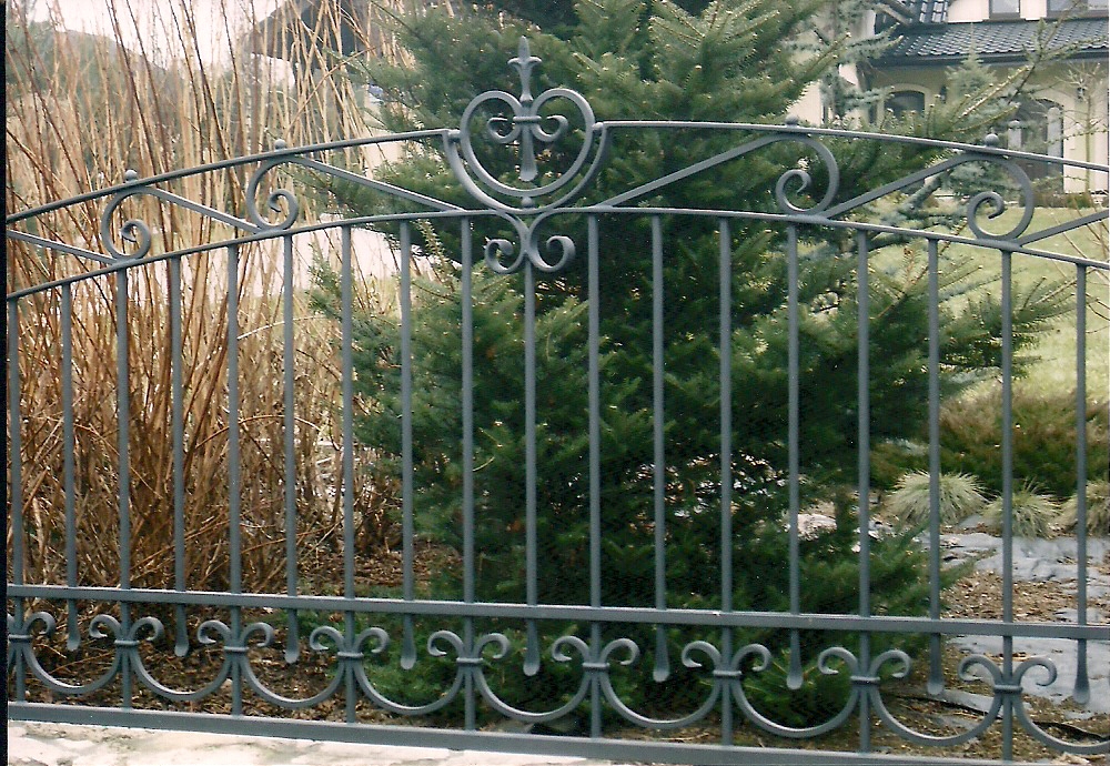 Kuta, metalowa brama ogrodzeniowa z dekoracyjnymi elementami w kształcie lilii i serca, na tle iglaka i trawy.