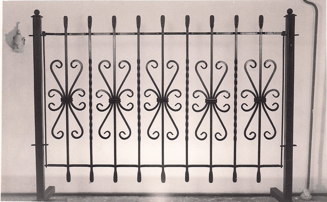 Czarno-białe zdjęcie fragmentu metalowej balustrady z ozdobnymi, symetrycznymi elementami w kształcie stylizowanych kwiatów i serc, zakończonych prostymi prętami.