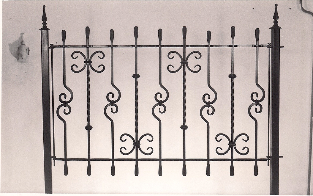 Kuta, metalowa brama ogrodzeniowa w stylu retro z ozdobnymi elementami w kształcie ślimaków i rozet na tle jasnej ściany.