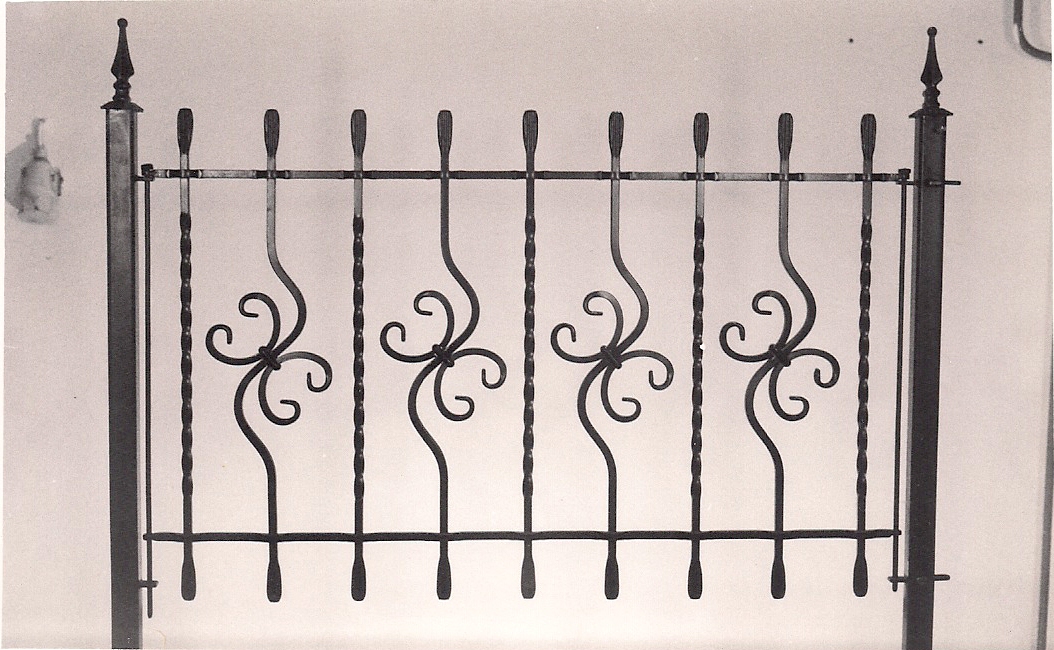 Kute, metalowe ogrodzenie z dekoracyjnymi elementami w kształcie czterolistnych koniczynek, zwieńczone ozdobnymi grotami, w czarno-białej tonacji.