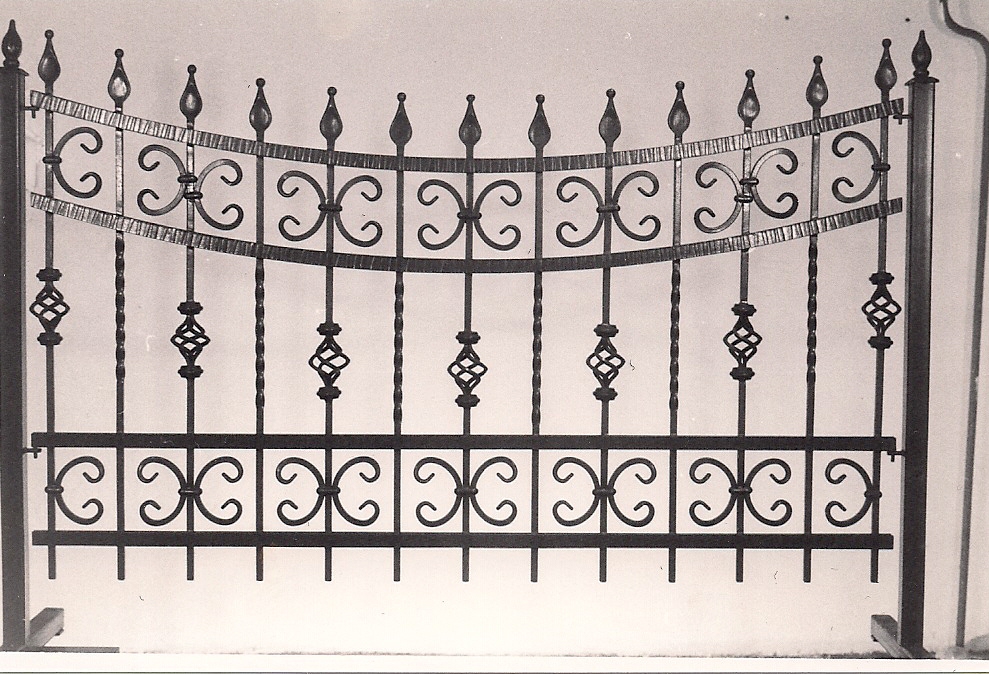 Czarno-białe zdjęcie fragmentu kutego ogrodzenia metalowego z ozdobnymi elementami w kształcie spiral i grotów.