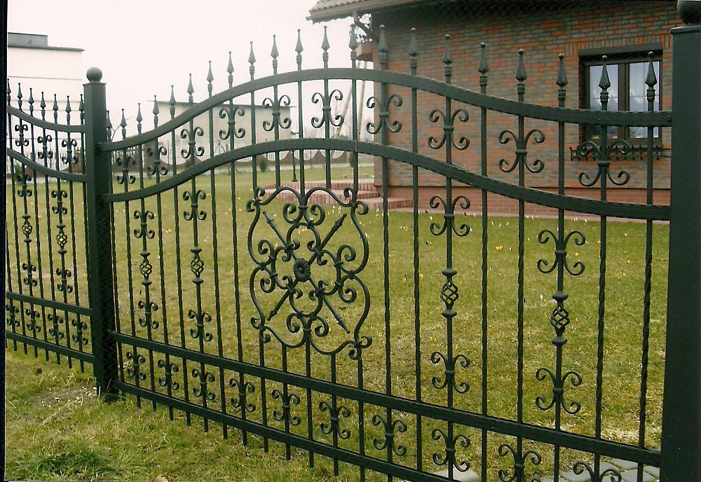 Kute, metalowe ogrodzenie z ozdobnymi elementami w kształcie fleur-de-lis i rozet, zabezpieczające posesję z trawnikiem i domem z cegły w tle.