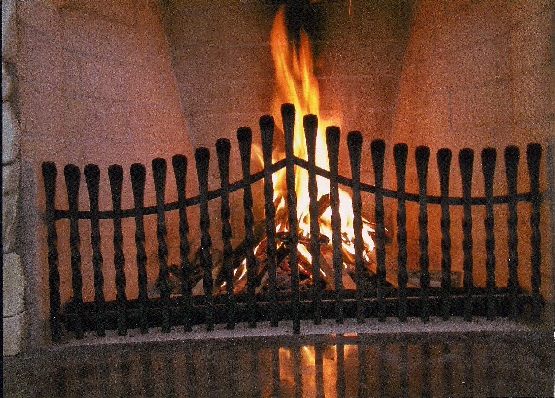 Kuty, czarny płotek kominkowy z pionowymi prętami i poziomą poprzeczką, chroniący palenisko z intensywnym ogniem w ceglanym kominku.