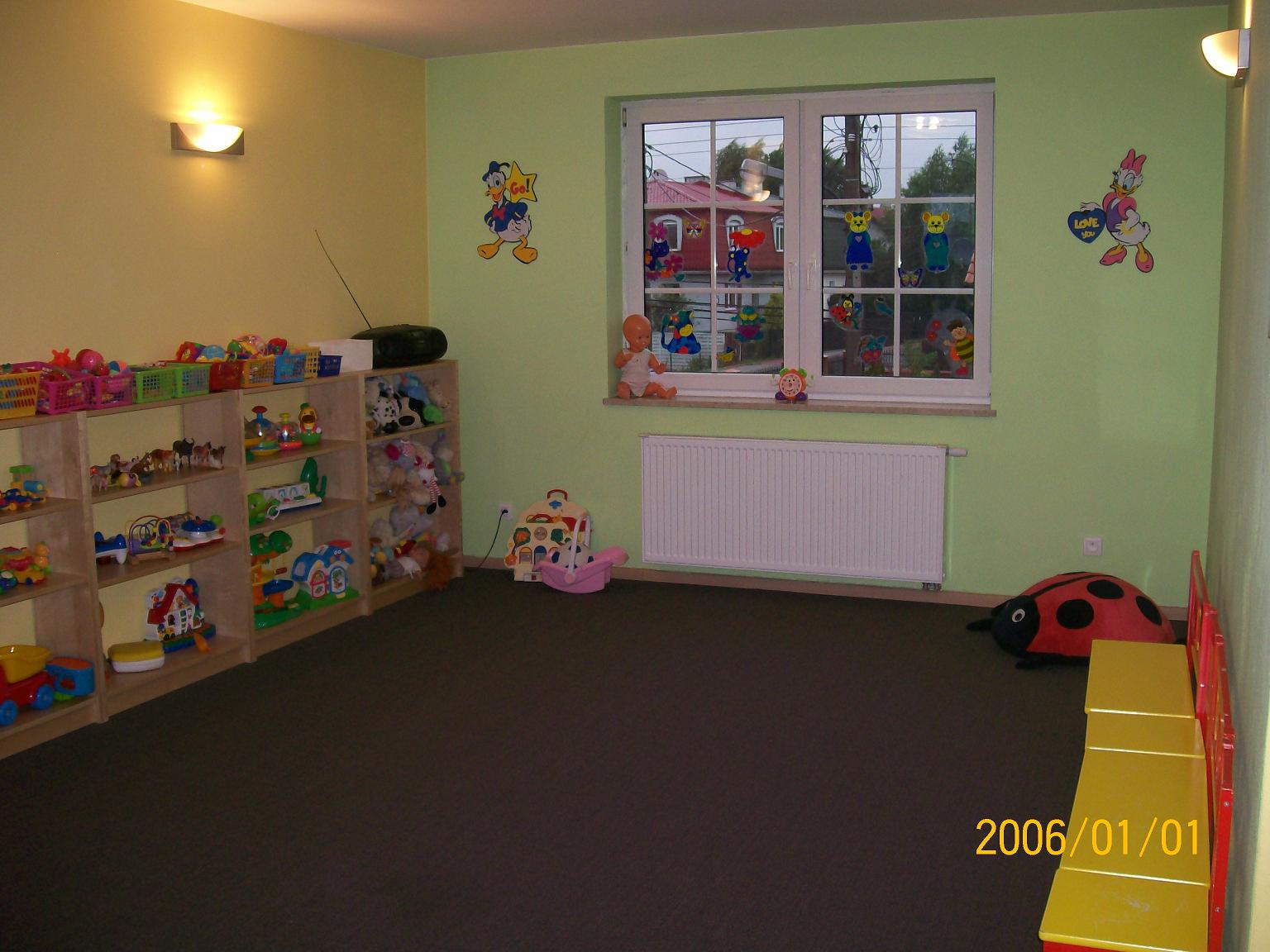 Wnętrze sali zabaw z regałami pełnymi zabawek, okno ozdobione naklejkami postaci z bajek, pluszowa biedronka na podłodze i mała zjeżdżalnia.