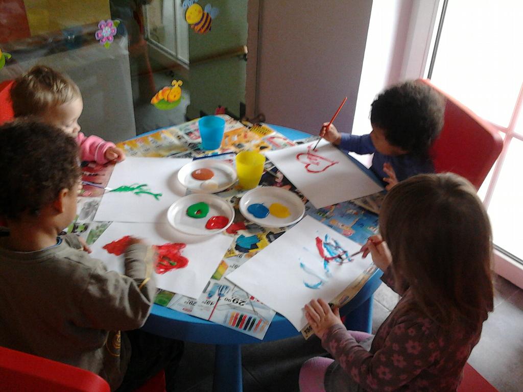 Czterech małych artystów przy niebieskim stole maluje farbami plakatowymi na białych kartkach, używając małych talerzyków jako palet z kolorowymi farbami; dekoracje pszczół i kwiatów na ścianie w tle.