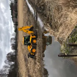 Żółta koparko-ładowarka JCB stojąca na brzegu cieku wodnego, odbicie maszyny w wodzie, w tle trzciny i drzewa, pochmurne niebo.