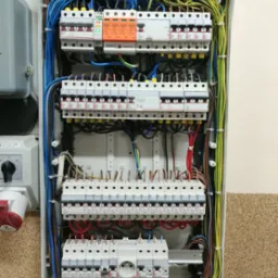 Otwarta skrzynka elektryczna z bezpiecznikami, przewodami w kolorach: niebieskim, ż&oacute;łto-zielonym, brązowym i czerwonym, oraz urządzeniami marki SIMTEC.