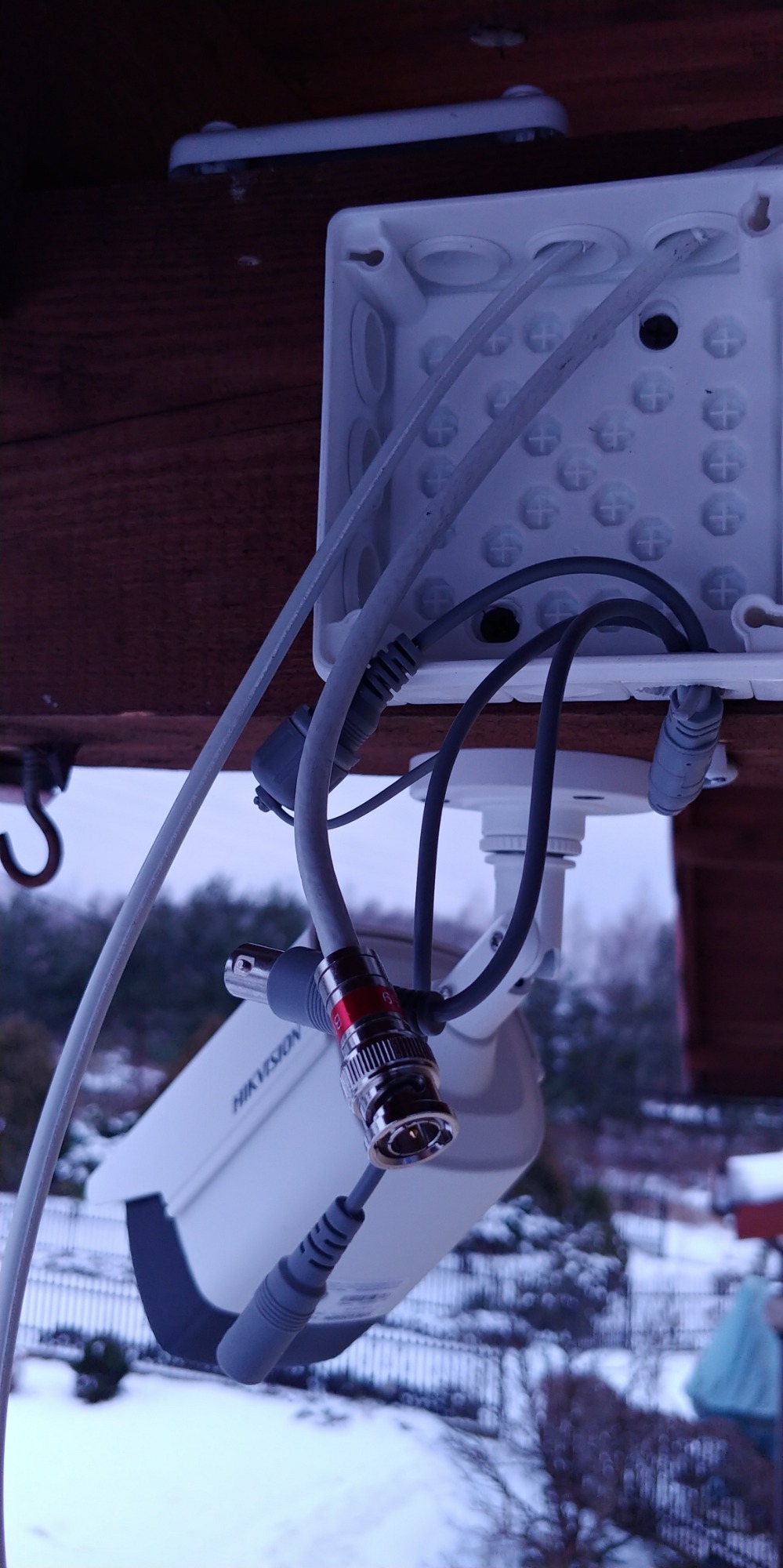 Kamera monitoringu Hikvision z widocznymi kablami i złączem BNC, zamontowana pod okapem w zimowej scenerii.