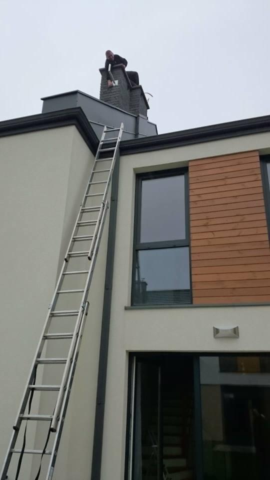 Mężczyzna na drabinie przy kominie na dachu domu, prawdopodobnie podczas czyszczenia lub inspekcji.