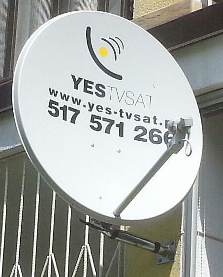 Biała antena satelitarna z logo YES TVSAT zamontowana na ścianie budynku. Widoczne numery telefonów i adres strony internetowej firmy.