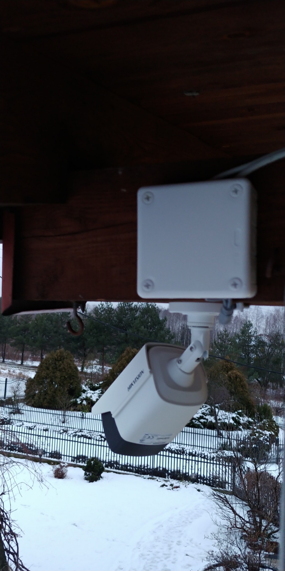 Zewnętrzna kamera monitoringu Hikvision zamontowana pod drewnianym zadaszeniem, widok na zasypany śniegiem ogród przez czarne metalowe ogrodzenie.