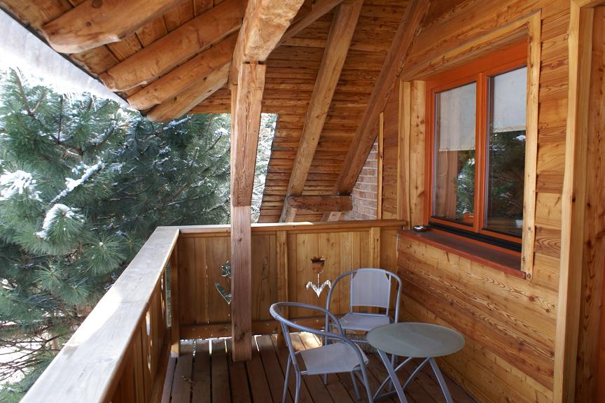 Drewniany balkon z widokiem na zasypane śniegiem drzewa, dwa krzesła i mały stolik. Konstrukcja dachu z widocznymi belkami. Okno z brązową ramą i częściowo zasłoniętą roletą.