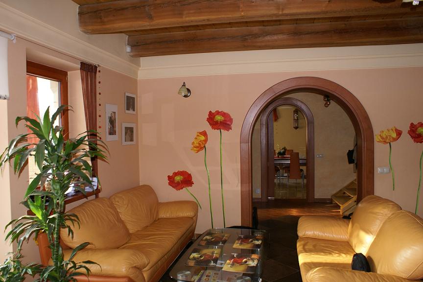 Jasny salon z dwoma skórzanymi kanapami, szklanym stolikiem kawowym, rośliną doniczkową przy oknie i dekoracyjnymi naklejkami w kształcie maków na ścianie, widok na jadalnię przez łukowate przejście.